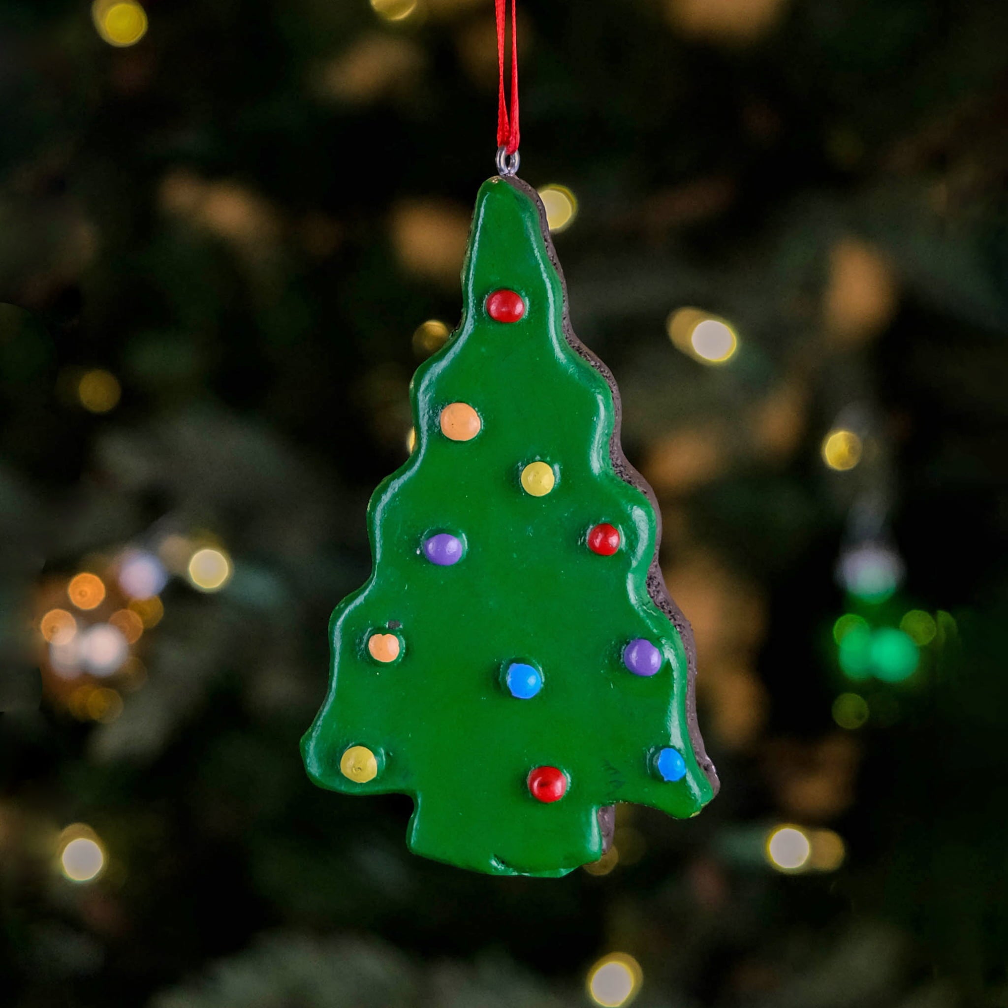 A Little Debbie® Christmas Tree Brownie® Ornament shaped like a green frosted Christmas tree brownie decorated with colorful candy pieces, hanging by a red ribbon in front of a softly lit Christmas tree background.