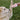 A light-colored dog raises its paw toward a plush, white Christmas Tree Cake dog toy embellished with red stripes and green sprinkles. A person’s hand holds the toy out in front of the dog, and they are outdoors on a grassy lawn.