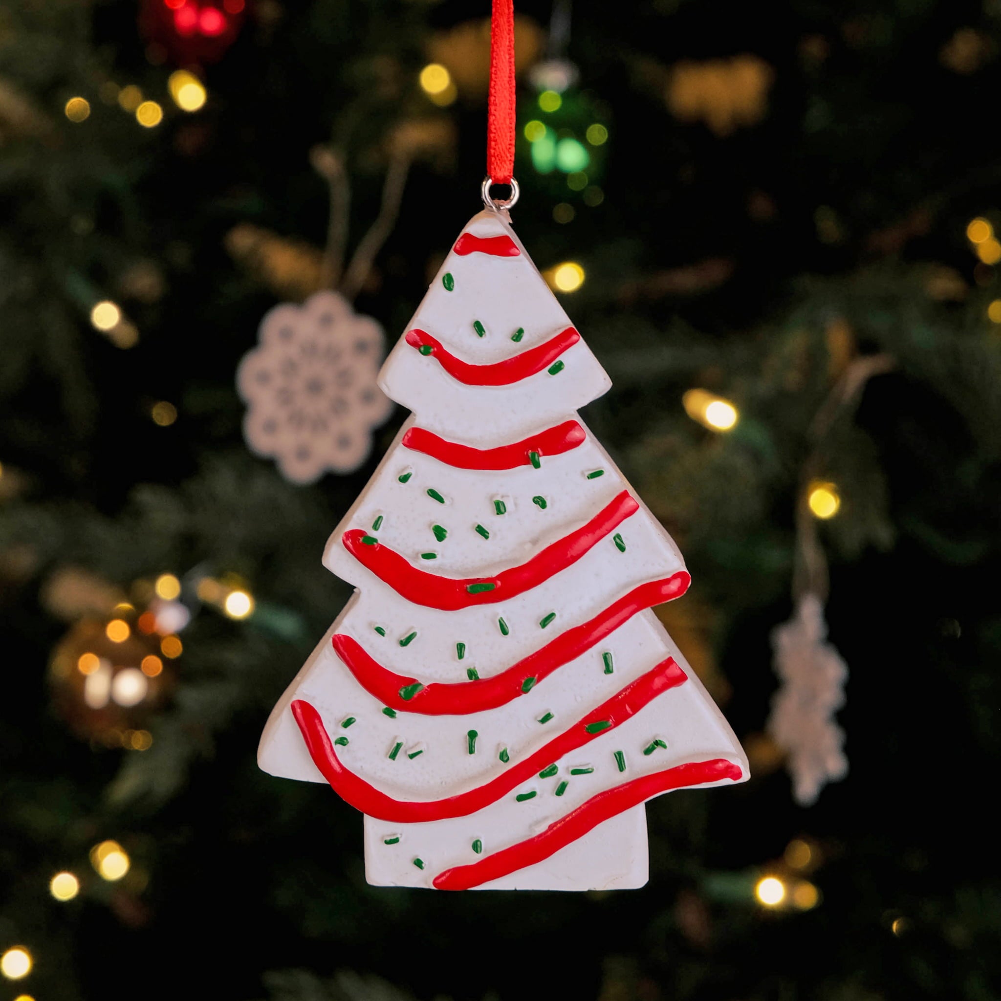 A Little Debbie Christmas Tree Cake® Ornament featuring white frosting with red icing stripes and green sprinkles, hanging by a red ribbon on a Christmas tree with twinkling lights in the background.