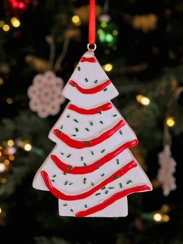 A Little Debbie Christmas Tree Cake® Ornament featuring white frosting with red icing stripes and green sprinkles, hanging by a red ribbon on a Christmas tree with twinkling lights in the background.