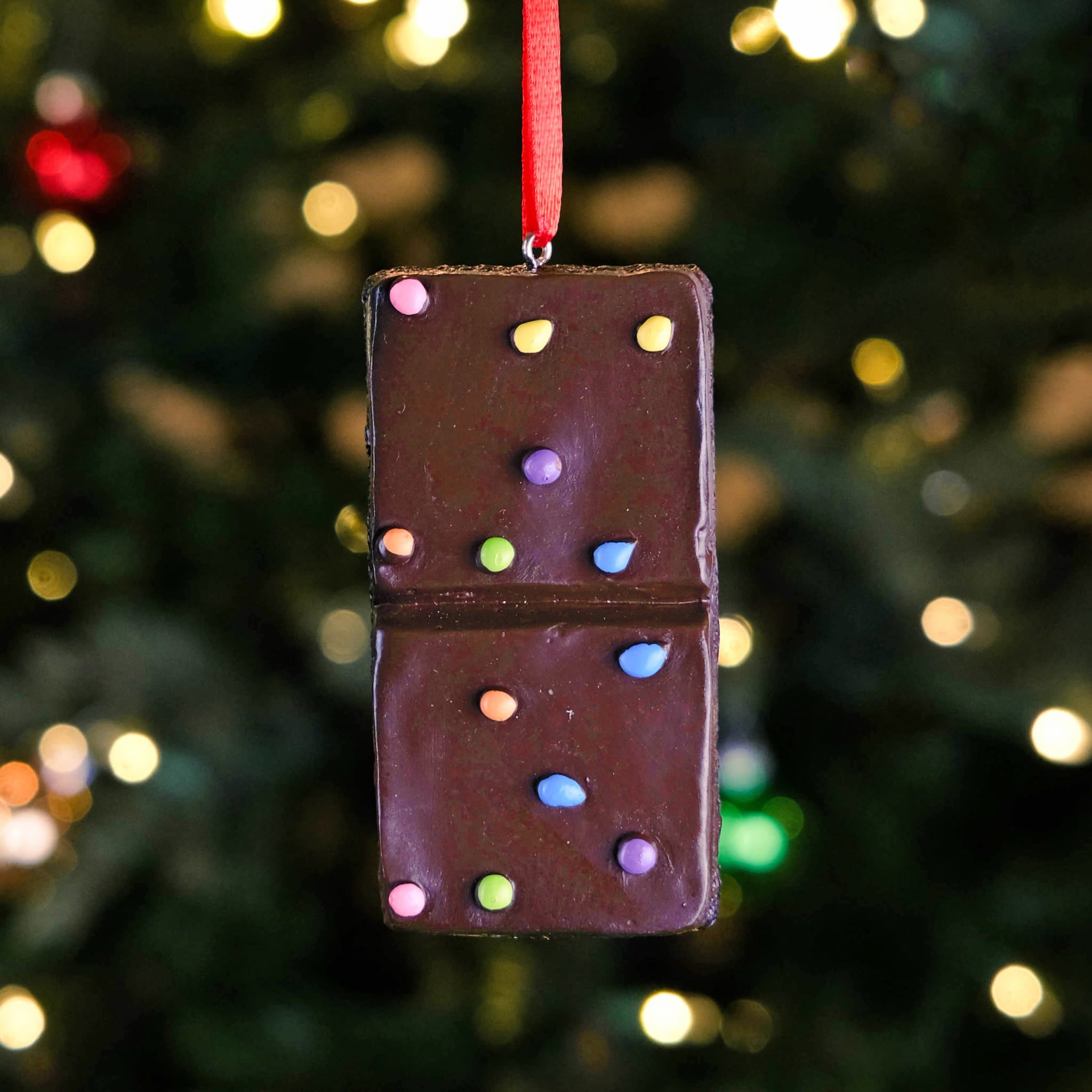 A Little Debbie®  Cosmic® Brownie Ornament hanging on a Christmas tree, featuring a realistic brownie design with colorful candy pieces and a red ribbon for hanging. Warm holiday lights softly blur in the background.
