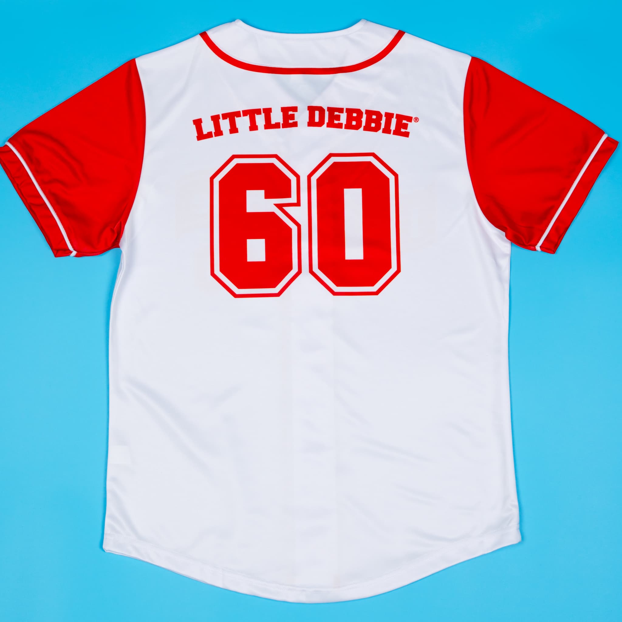 A white and red Little Debbie® baseball jersey displayed flat on a blue background. The jersey features red sleeves, red piping, and bold red lettering across the front that reads “Little Debbie” along with the number “60” to represent the brand’s founding year.