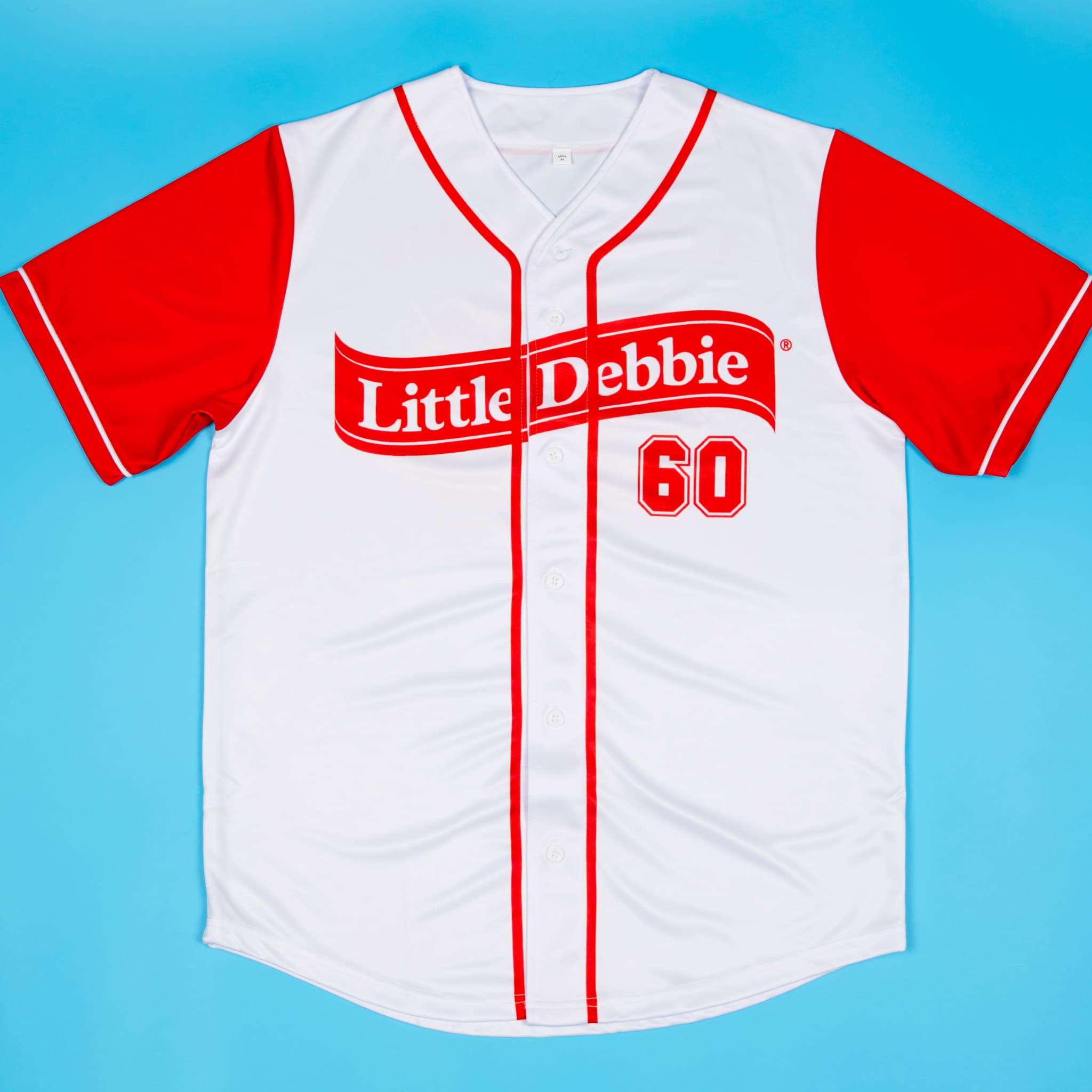 A white and red Little Debbie® baseball jersey displayed flat on a blue background. The jersey features red sleeves, red piping, and bold red lettering across the front that reads “Little Debbie” along with the number “60” to represent the brand’s founding year.