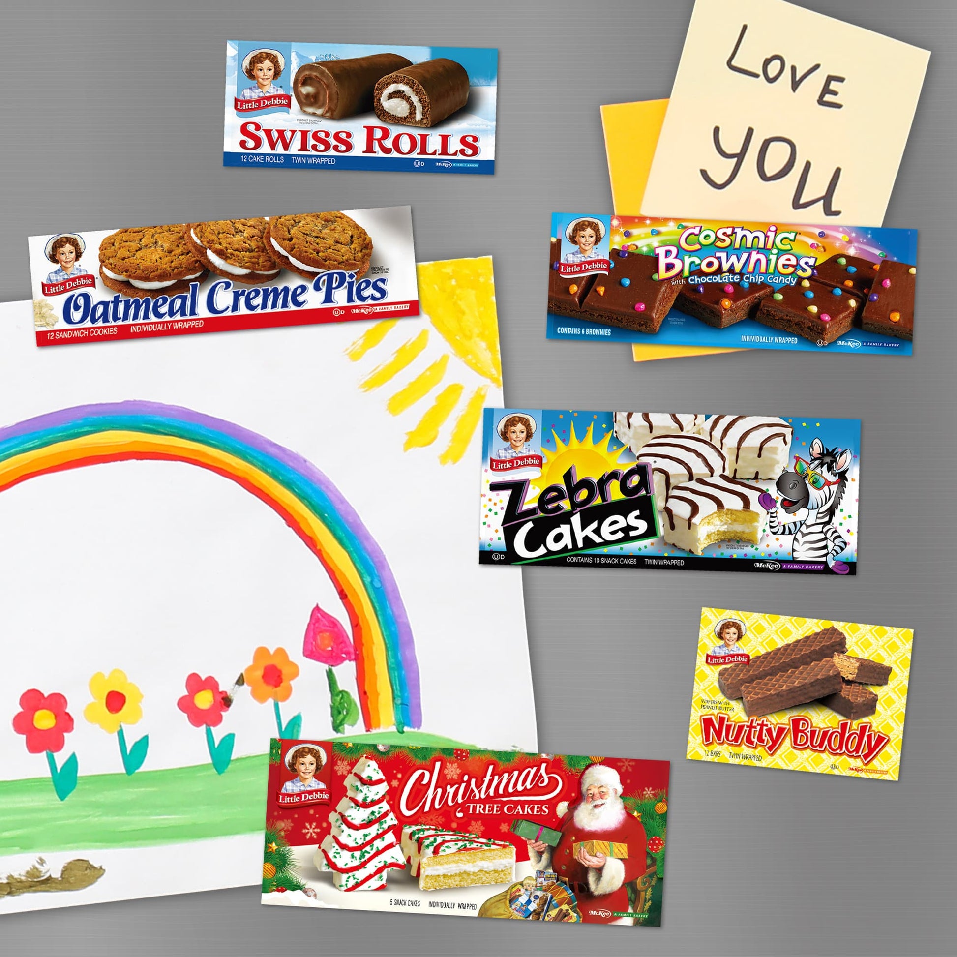 Collection of Little Debbie snack carton magnets arranged on a silver background, featuring popular treats. The set includes Swiss Rolls, Oatmeal Creme Pies, Cosmic Brownies, Zebra Cakes, Christmas Tree Cakes, and Nutty Buddy. Each magnet is a colorful replica of the snack's original packaging. A hand-drawn childlike illustration of a rainbow with flowers and a 'Love You' note on yellow paper are also part of the playful display.