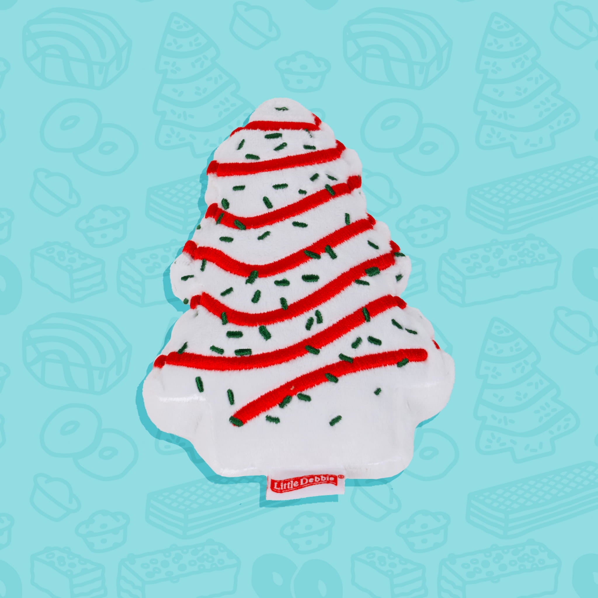 A Little Debbie Christmas Tree Cake dog toy shaped like the classic holiday treat. The toy is white with red icing stripes and green sprinkle details, resembling the festive design of the Christmas Tree Cake snack. A small red Little Debbie logo tag is attached at the bottom. The background is light blue with faint outlines of various Little Debbie snacks.