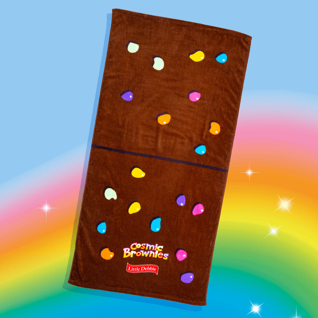 A Little Debbie® Cosmic Brownie beach towel designed to resemble the popular Cosmic® Brownie snack. The towel is chocolate brown with colorful, candy-like sprinkle shapes scattered across, similar to the topping on Cosmic® Brownies. The "Cosmic® Brownies" logo is printed in vibrant colors near the bottom, along with a red Little Debbie logo. The towel is displayed on a rainbow-colored background with star-like sparkles.