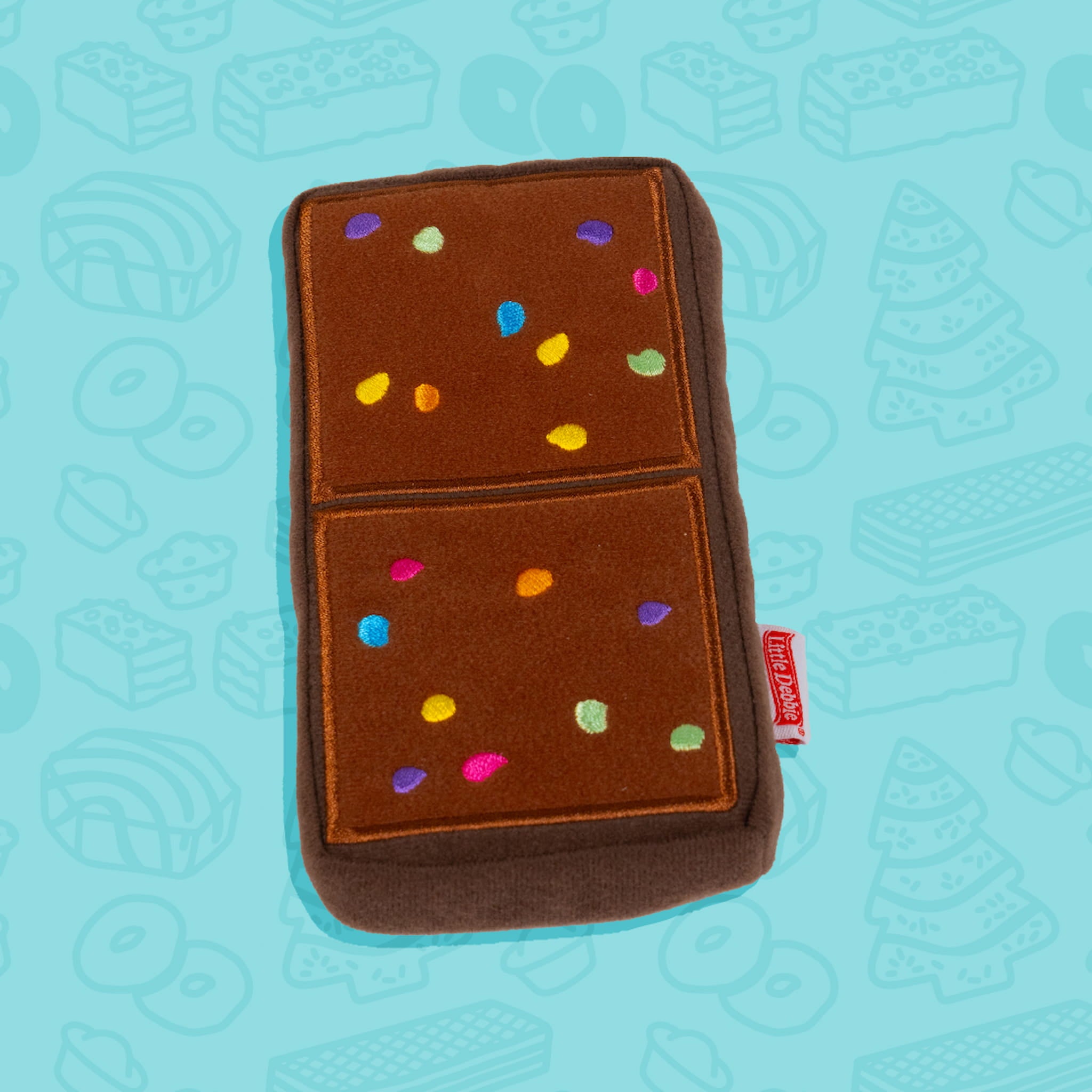 A Little Debbie Cosmic Brownie dog toy shaped like the iconic Cosmic Brownie snack. The toy is rectangular with a chocolate-brown color, divided into two sections, each decorated with colorful sprinkle-like dots in blue, green, yellow, pink, purple, and orange. A small red Little Debbie logo tag is attached to the side. The background is light blue with faint outlines of various Little Debbie snacks.