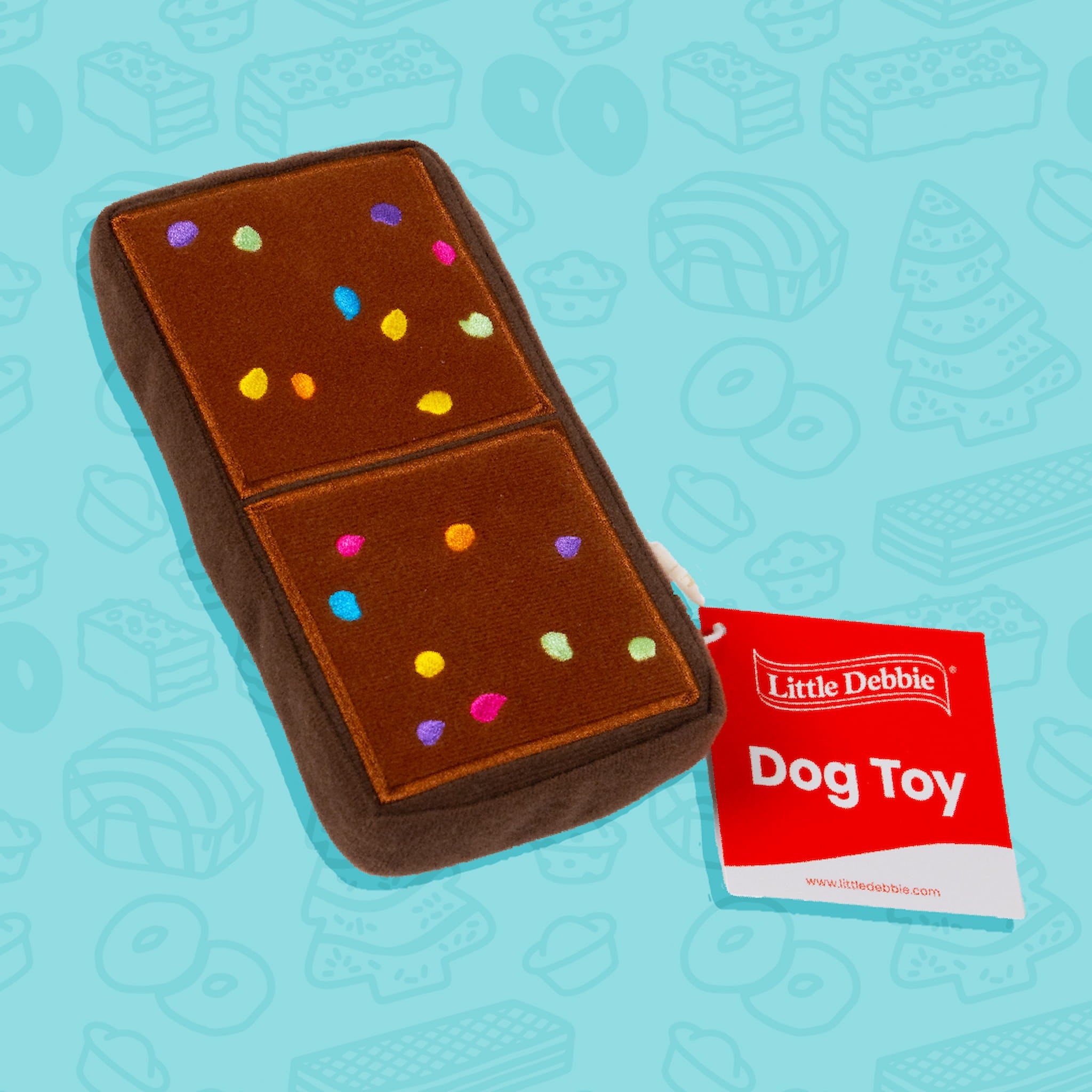 A Little Debbie Cosmic Brownie dog toy designed to resemble the classic Cosmic Brownie snack. The toy is brown with a rectangular shape divided into two sections, each decorated with colorful sprinkle-like dots in yellow, blue, green, purple, and pink. A red tag attached to the toy reads "Little Debbie Dog Toy." The background is light blue with faint outlines of various Little Debbie snacks.