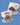 A Little Debbie gingham mug featuring a charming snack-themed design. Each white mug is accented with a light blue gingham border near the bottom and a matching light blue handle. The design showcases illustrations of Little Debbie snacks, including Oatmeal Creme Pies, Nutty Buddy Bars, Cosmic Brownies, and Swiss Rolls, with their names written in cursive. The red Little Debbie logo is prominently displayed near the top of the mug. 