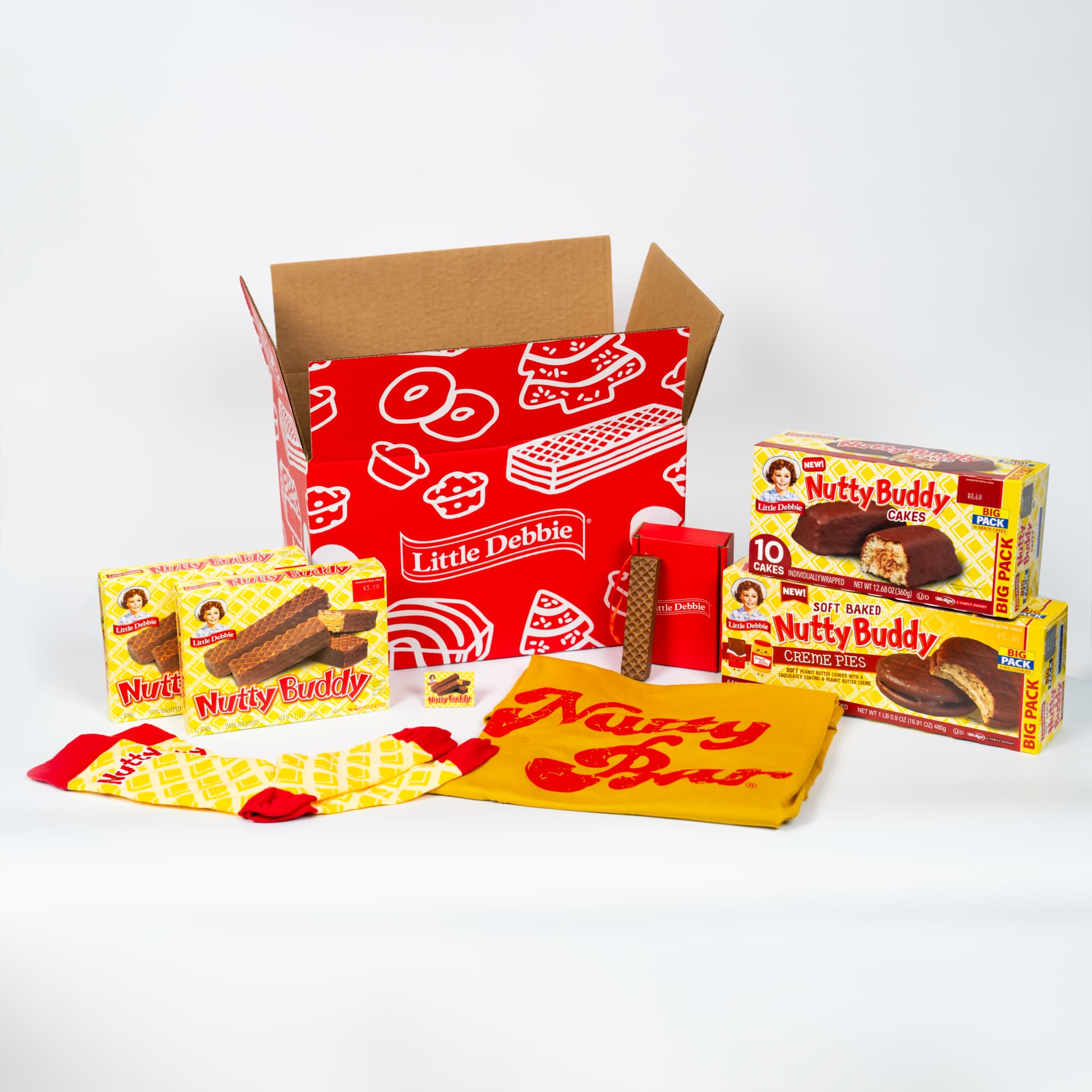 A Nutty Buddy™ themed gift box display featuring two cartons of classic Nutty Buddy bars, one carton of Nutty Buddy Cakes, one carton of Nutty Buddy Creme Pies, a yellow Nutty Buddy t-shirt, red-and-yellow Nutty Buddy socks, a Nutty Buddy carton magnet, an ornament, and a bright red Little Debbie Treat Trove shipping box in the background. All items are arranged on a white surface with a clean, vibrant presentation.