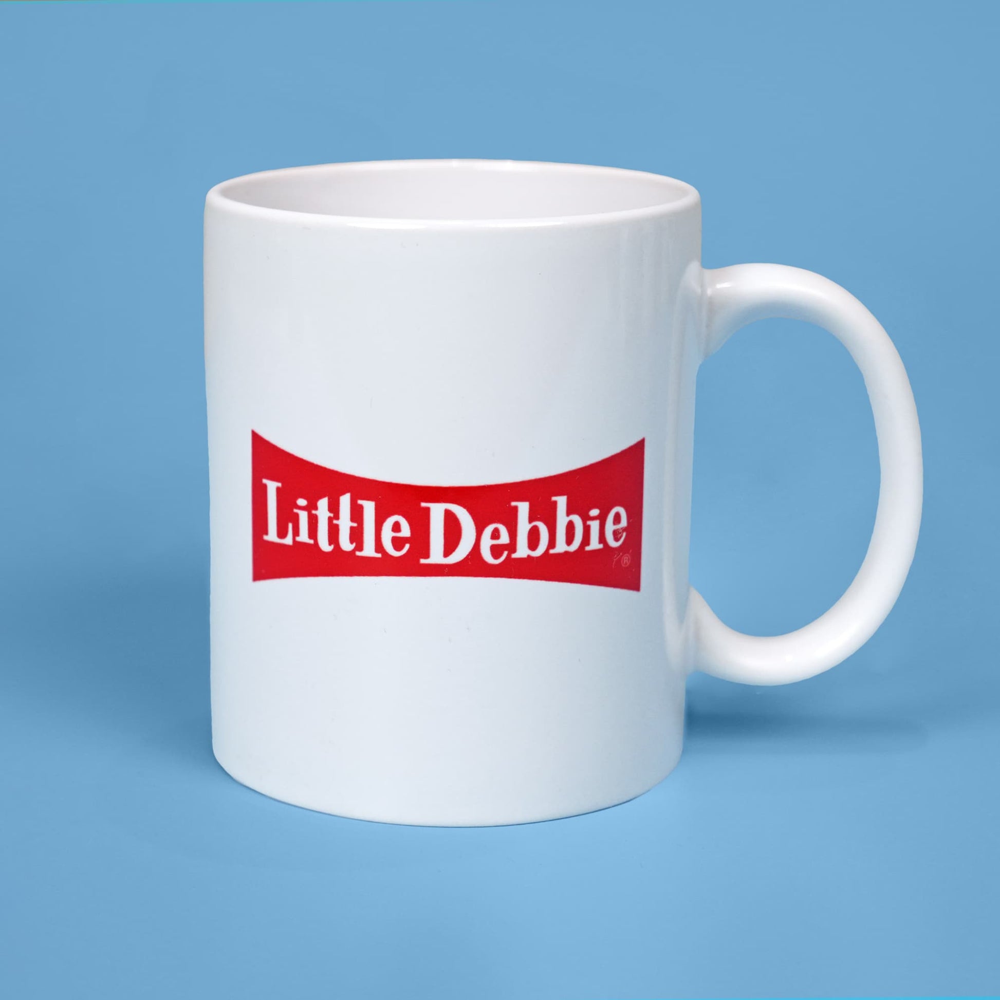 White ceramic mug with a red banner featuring the 'Little Debbie' logo in white letters, set against a light blue background. The mug has a simple, clean design with a classic brand logo, emphasizing the red and white color scheme.