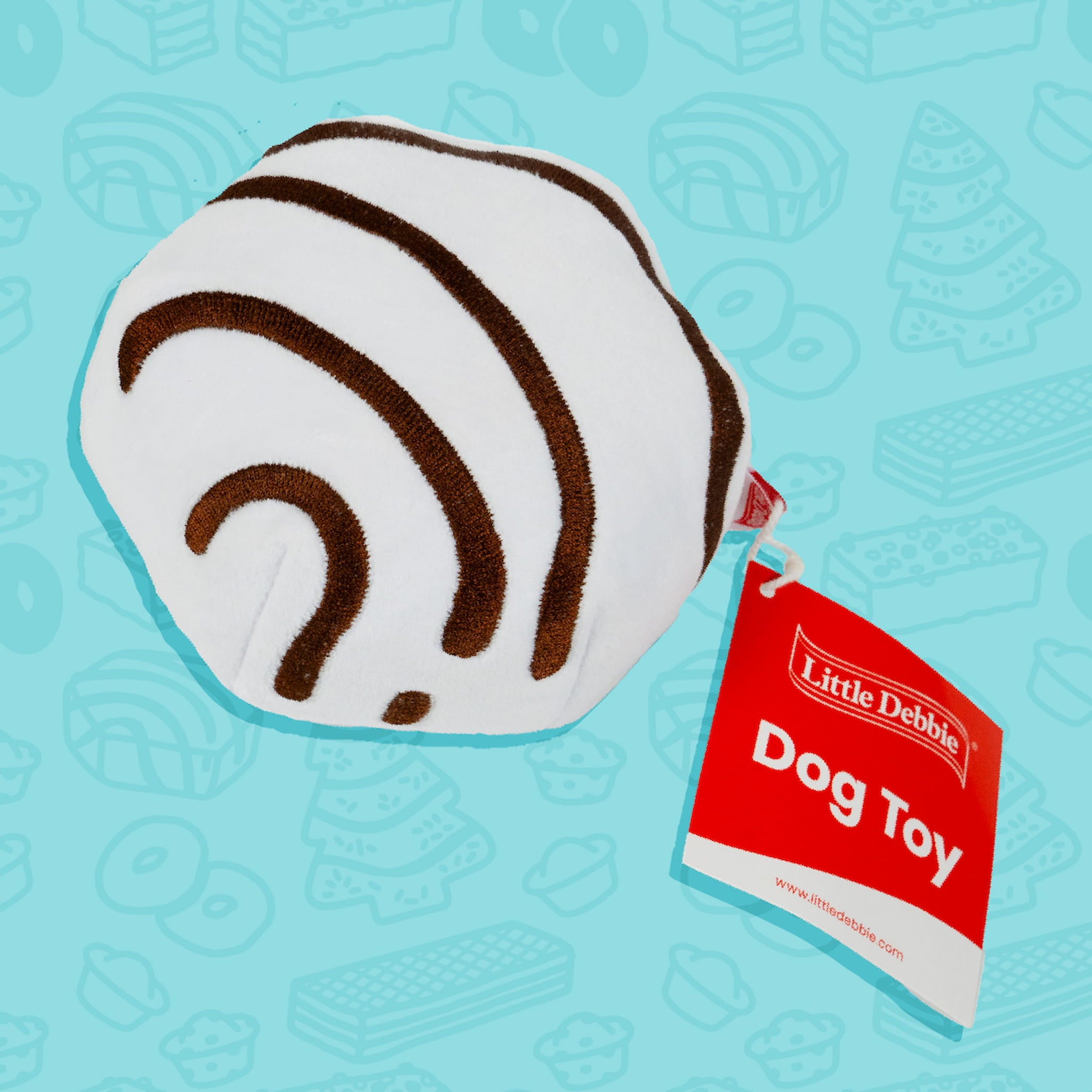 A Little Debbie Zebra Cake dog toy designed to resemble the iconic Zebra Cake snack. The toy is round with a white base and features the signature brown zebra-like stripes. A red tag attached to the toy reads "Little Debbie Dog Toy." The background is light blue with faint outlines of various Little Debbie snacks.