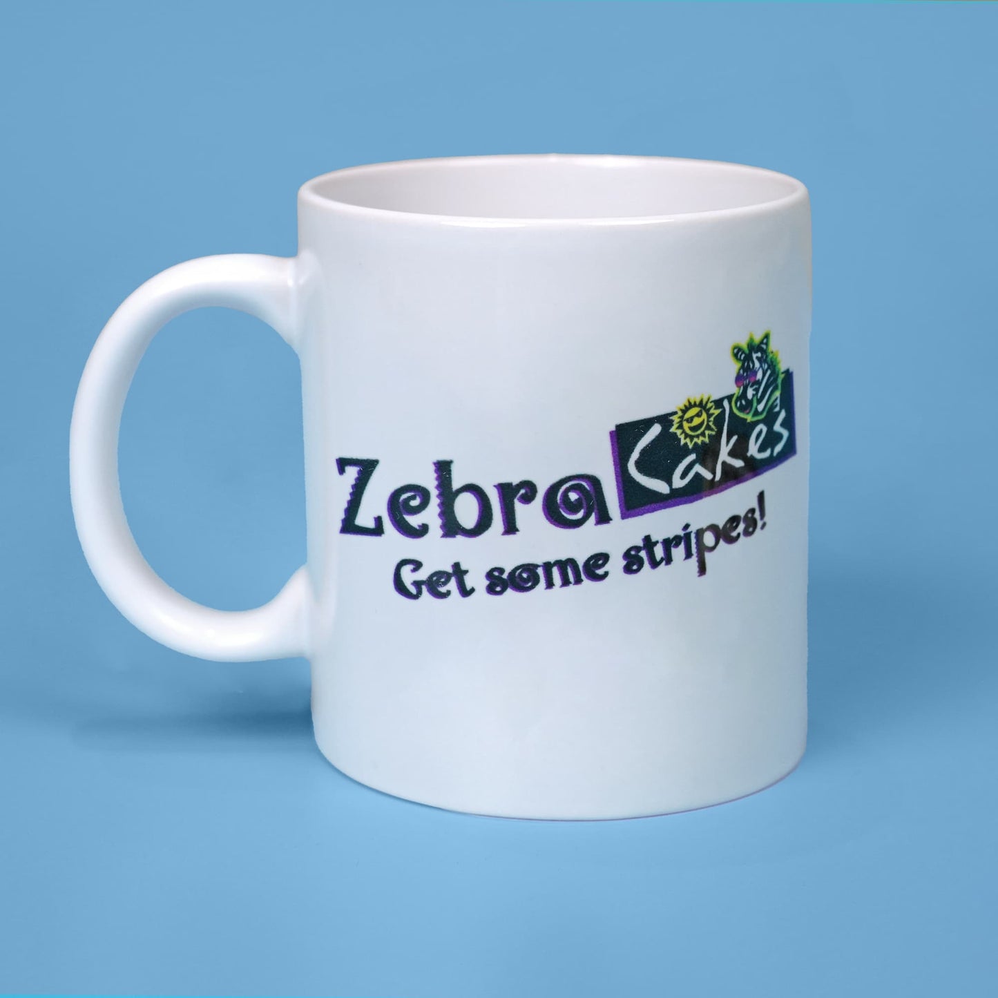 White ceramic mug with the 'Zebra Cakes' logo in purple letters and the slogan 'Get some stripes!' on it. Next to the text, there is a vibrant illustration of a cartoon zebra with a sun, both in purple, black, and yellow. The background of the mug is a soft blue.