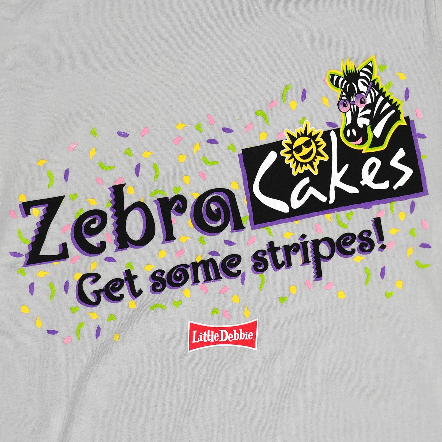 Close-up of the graphic on a light gray t-shirt, displaying the 'Zebra Cakes' logo in bold black and purple letters, with the phrase 'Get some stripes!' underneath. Adjacent to the text is a colorful illustration of a cartoon zebra and a sun in yellow and black. The background of the graphic is scattered with small paint splatters in purple, green, and yellow. A small red Little Debbie logo is visible in the lower right of the design.