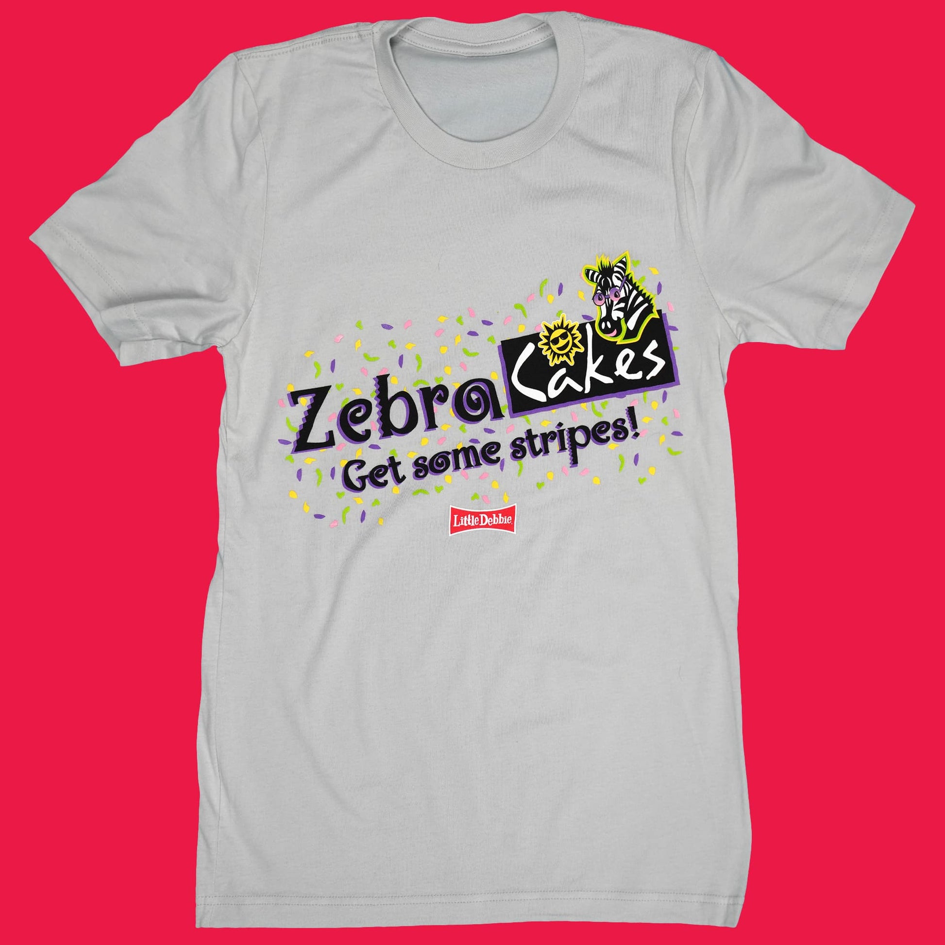 Light gray t-shirt featuring a vibrant design for Little Debbie Zebra Cakes. The logo in black and purple reads 'Zebra Cakes' with the slogan 'Get some stripes!' underneath in yellow. There's a playful depiction of a cartoon zebra and a sun, both in yellow and black, to the right of the text. The background is speckled with small, colorful paint splatters in purple and yellow. A small red Little Debbie logo is positioned below the main design.