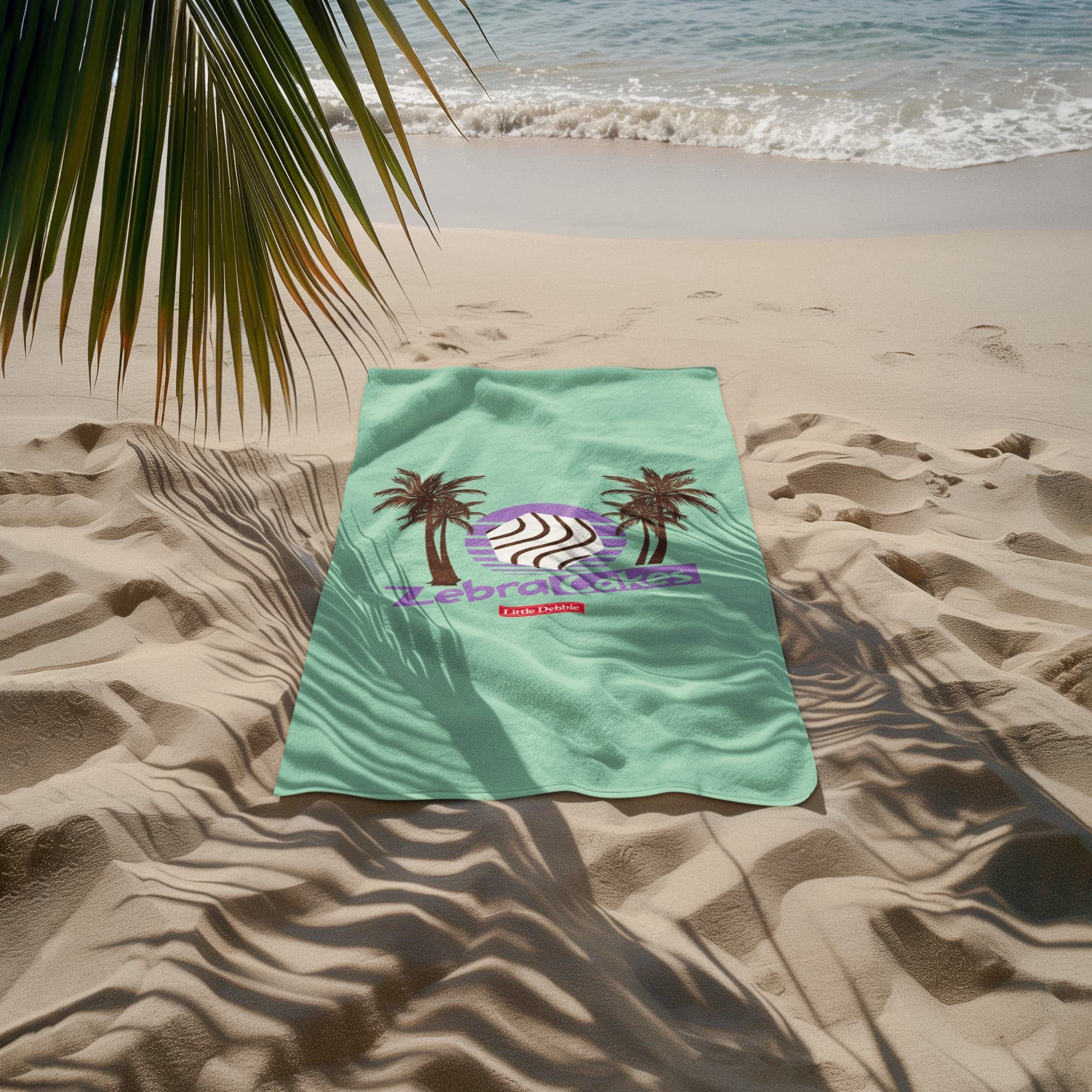 A Little Debbie® Zebra Cakes palm beach towel spread out on sandy beach with gentle waves in the background. The towel is teal with a design featuring two black palm trees framing a stylized Zebra Cake in black-and-white stripes against a purple circle. Below, "Zebra Cakes" is written in bold purple, with the red Little Debbie® logo underneath. Palm fronds cast natural shadows across the towel and sand, enhancing the tropical beach setting.