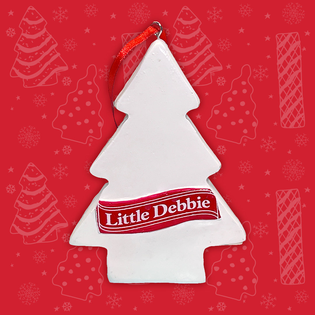The back side of the Little Debbie Christmas Tree Cake ornament, showcasing a clean white surface with the 'Little Debbie' logo across a red banner. The ornament hangs by a festive red ribbon, set against a red background with subtle snowflake and Christmas Tree Cake and Brownie silhouettes.