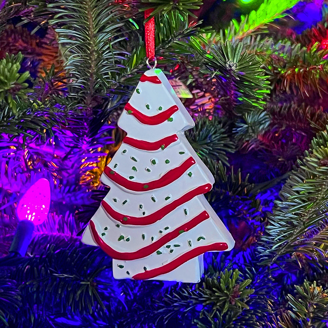 The Little Debbie Christmas Tree Cake ornament is festively hung on a Christmas tree, with its white icing and red garland design vivid against the dark green branches. Multi-colored lights, including a prominent purple bulb, illuminate the ornament, enhancing its holiday charm.