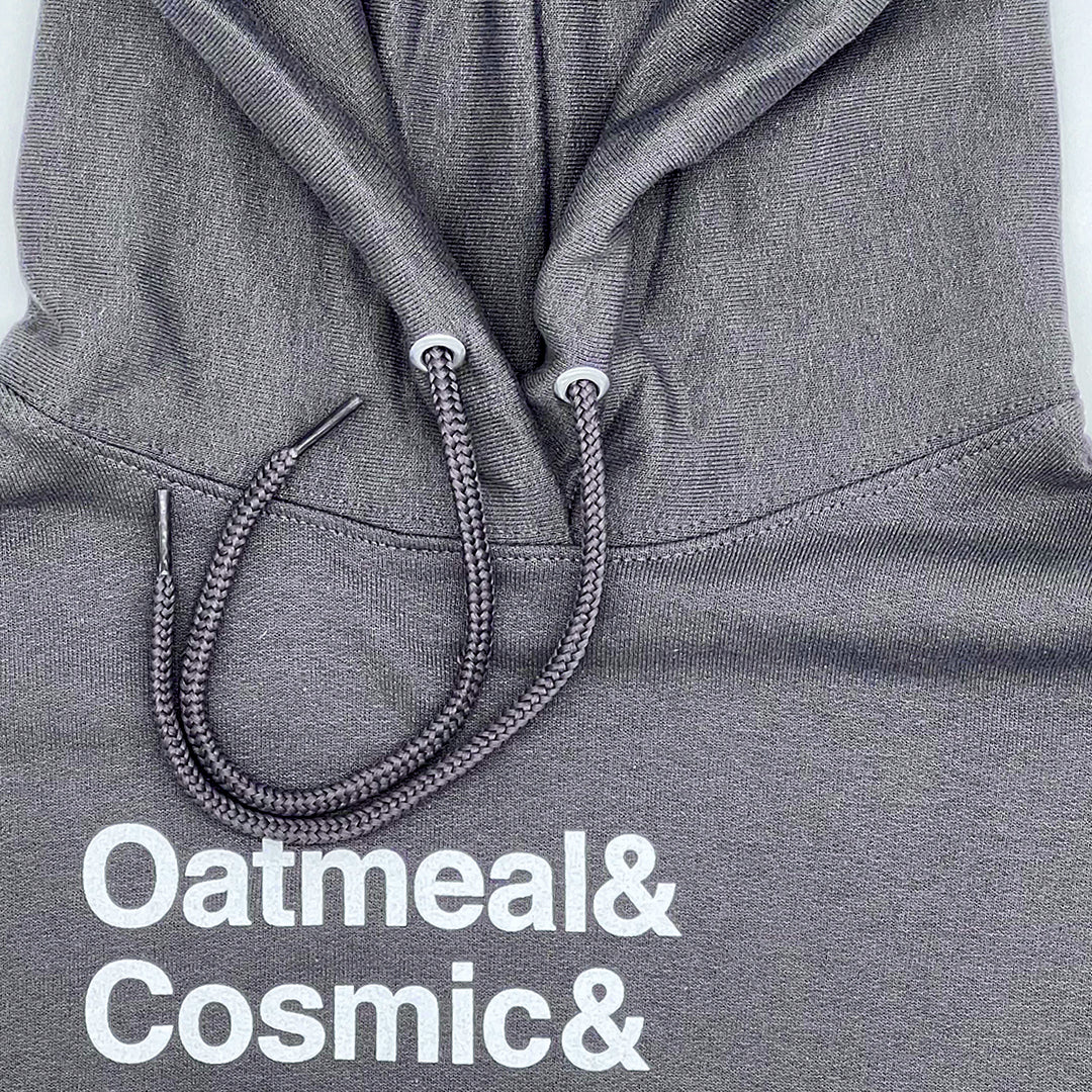 Close-up view of the top part of a Little Debbie Fan Favorites gray hoodie. The fabric displays a finely knit texture, and the drawstrings are braided with metal aglets. Just below the drawstrings, white text reads 'Oatmeal& Cosmic&'. 