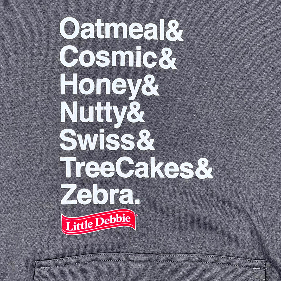 Close-up of a Little Debbie Fan Favorites hoodie's gray fabric with white text listing various snack names in a vertical format: 'Oatmeal& Cosmic& Honey& Nutty& Swiss& TreeCakes& Zebra.' Below the list, a Little Debbie red ribbon logo is prominently displayed. The bottom edge of the fabric appears to be a hem or pocket seam.