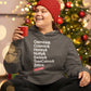 Cheerful woman wearing a Little Debbie Fan Favorites gray hoodie with white text listing snack names like 'Oatmeal, Cosmic, Honey, Nutty, Swiss, TreeCakes and Zebra', with a Little Debbie red ribbon logo at the bottom. She is holding a red mug and wearing a red knit hat, sitting in front of a brightly lit Christmas tree adorned with ornaments.