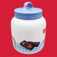 The image shows a Little Debbie Collectible Cookie Jar on a red background, featuring a blue gingham design at the base. The jar is white and has a depiction of Cosmic Brownies, complete with colorful candy pieces on top. The text 'Cosmic Brownies' is written in a fun, casual script next to the image. The jar is topped with a blue lid that has a simple loop handle.