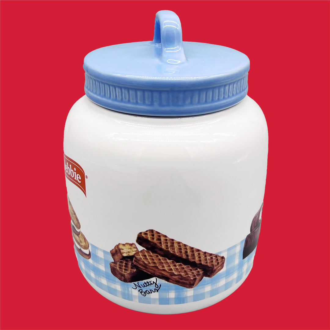 A Little Debbie collectible gingham cookie jar is depicted against a red background. The cylindrical jar is white with a blue checkered gingham pattern along the bottom. It features an image of Little Debbie Nutty Buddy with the text 'Nutty Bars' in a cursive script. The jar has a prominent blue lid with a rounded handle on top, signaling easy accessibility. It bears the Little Debbie logo in red.