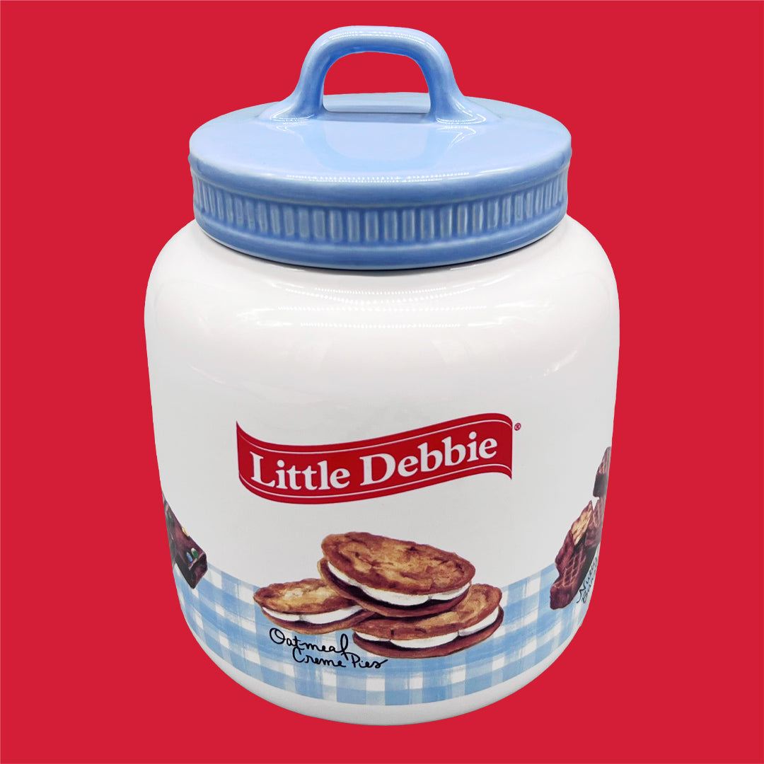 A collectible Little Debbie® cookie jar featuring an illustration of Oatmeal Creme Pies. The jar is white with a blue gingham check pattern at the bottom. The lid is blue with a convenient handle. The brand's logo is prominently displayed at the center.