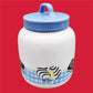 Featured is a Little Debbie collectible cookie jar with a blue gingham pattern at its base, set against a vibrant red background. The jar is predominantly white and showcases an image of a Zebra Cake, characterized by its distinctive white icing with fudge stripes. The words 'Zebra Cakes' are whimsically written below the illustration. Completing the design, the jar's blue lid has a convenient loop handle, enhancing the overall vintage charm of the piece.