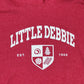 Close-up of a red fabric texture featuring the printed text 'LITTLE DEBBIE' in bold white letters. Below it, there's a shield emblem displaying various Little Debbie products, including a Swiss Cake swirl, a Zebra Cake, a Christmas Tree Cake, and pair of Nutty Buddys. The words 'EST. 1960' are printed beside the emblem.