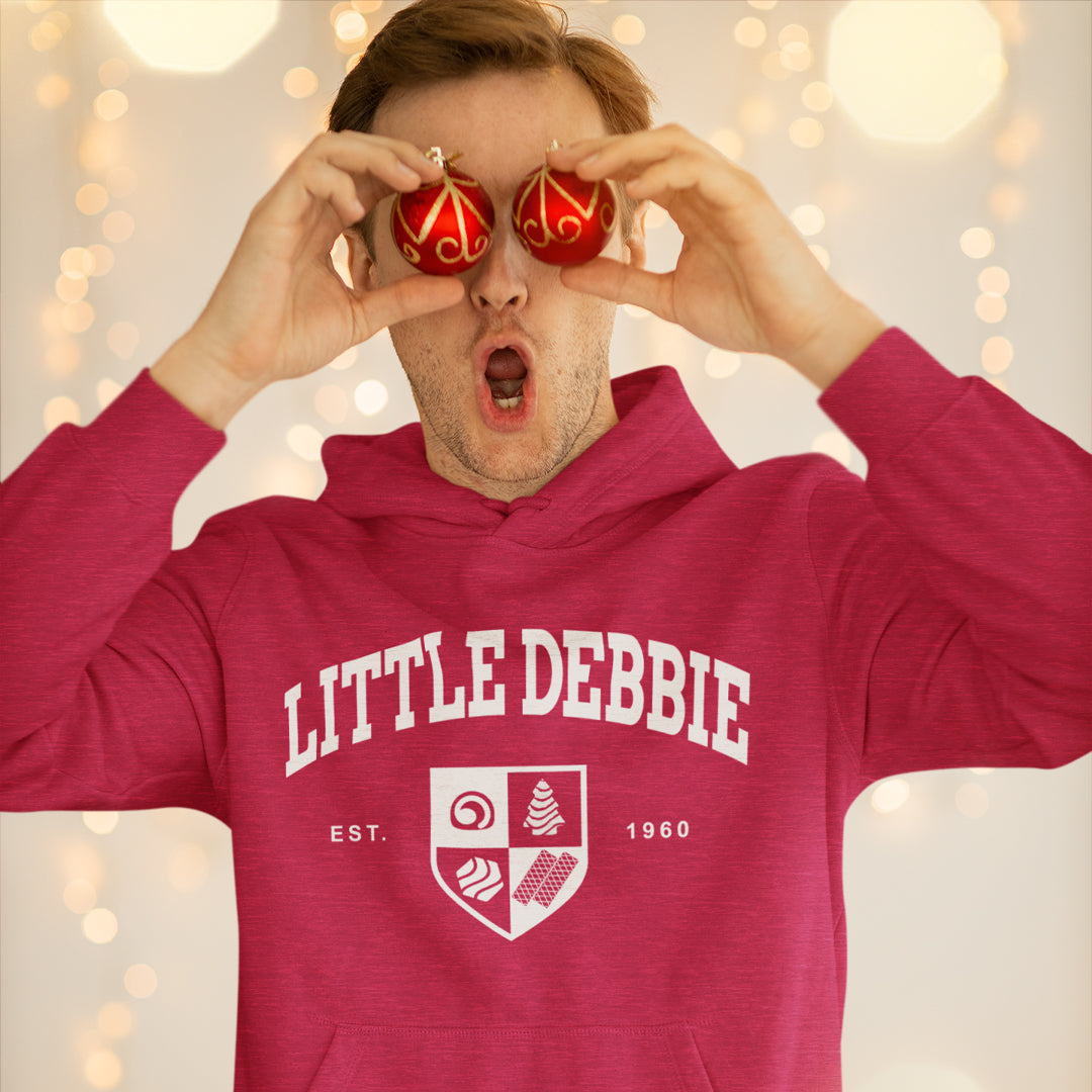 Man in a red 'LITTLE DEBBIE' hoodie with a logo reading 'EST. 1960' holds up two red and gold Christmas ornaments in front of his eyes, making a surprised expression. The background features soft bokeh lights.