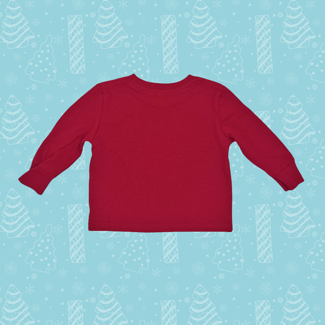 A red Little Debbie® Christmas Tree Cake Taste Tester toddler shirt with long sleeves is centered on a light blue background patterned with white Christmas trees and snowflakes. The shirt is plain, without any visible designs or text, and is laid out flat to show the shape and size suitable for a toddler. 