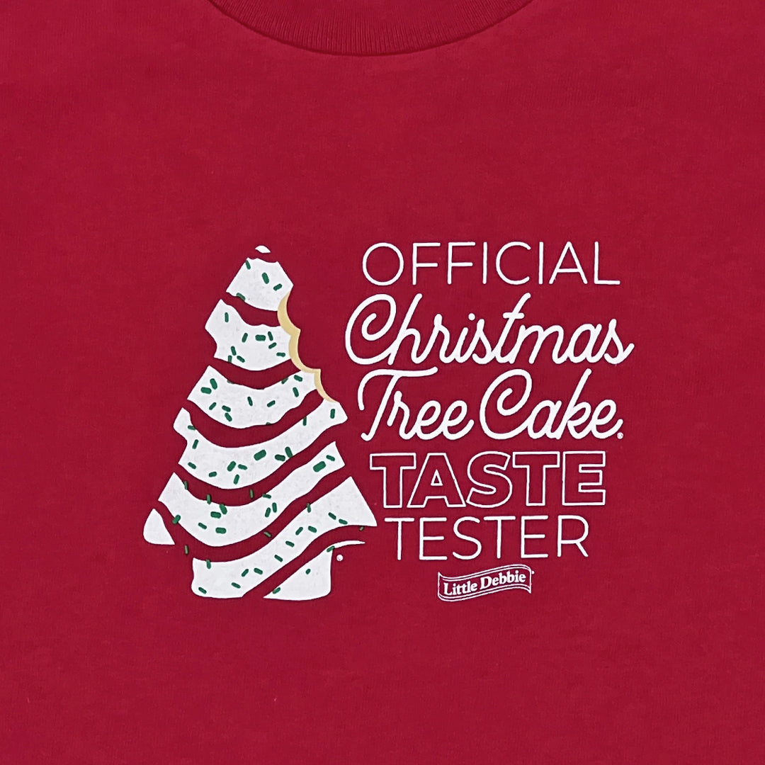 Close-up of a red toddler shirt featuring a white graphic of a Little Debbie® Christmas Tree Cake on the left side. Adjacent to the graphic, in white text, it reads 'OFFICIAL Christmas Tree Cake TASTE TESTER' with the Little Debbie® logo situated at the bottom right of the text. The shirt's neckline is round, and the background of the image is a solid red matching the shirt.