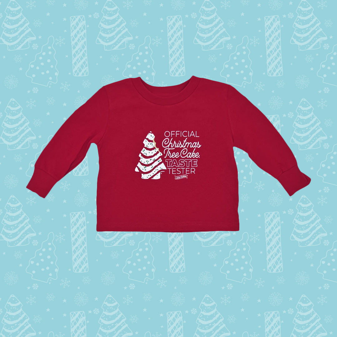 A red Little Debbie® Christmas Tree Cake Taste Tester Toddler Shirt with long sleeves, laid flat against a light blue background decorated with white Christmas tree and snowflake patterns. The shirt features a white printed design of a Christmas tree cake on the left side with the words 'OFFICIAL Christmas Tree Cake TASTE TESTER' written around it in a festive font, including the Little Debbie® logo at the bottom of the text.