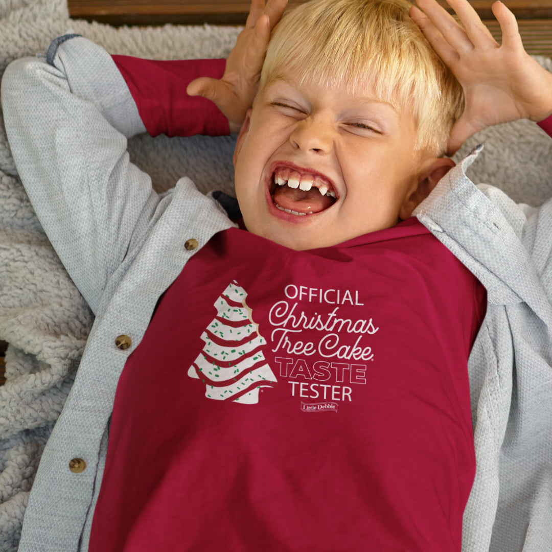 A joyful toddler is lying on their back with their hands playfully on their head, wearing a red Little Debbie® Christmas Tree Cake Taste Tester Toddler Shirt with the words 'OFFICIAL Christmas Tree Cake TASTE TESTER' printed on the front. The shirt also features a graphic of a Little Debbie® Christmas Tree Cake and the Little Debbie® logo. The child is also wearing a light gray button-up shirt over the red shirt and has a bright, excited expression with their eyes closed and mouth open in a wide smile.