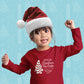 A smiling toddler with a Santa hat is raising one fist in excitement, wearing a red long-sleeve Little Debbie® Christmas Tree Cake Taste Tester Toddler Shirt that reads 'OFFICIAL Christmas Tree Cake TASTE TESTER' with a graphic of a Little Debbie® Christmas Tree Cake. The background is light blue with a festive pattern of white Christmas trees and snowflakes.