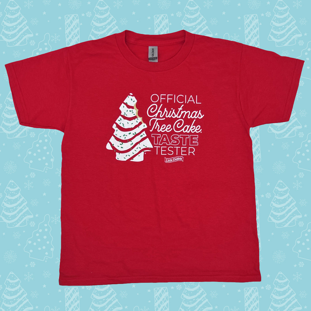 A red Little Debbie® Christmas Tree Cake Taste Tester Youth Shirt is displayed flat against a light blue background decorated with white illustrations of Christmas trees and snowflakes. The shirt features a printed graphic of a white Christmas Tree Cake on the left side, and white text on the right side reading 'OFFICIAL Christmas Tree Cake TASTE TESTER', with the Little Debbie® logo just below. The T-shirt has a classic crew neckline and short sleeves.