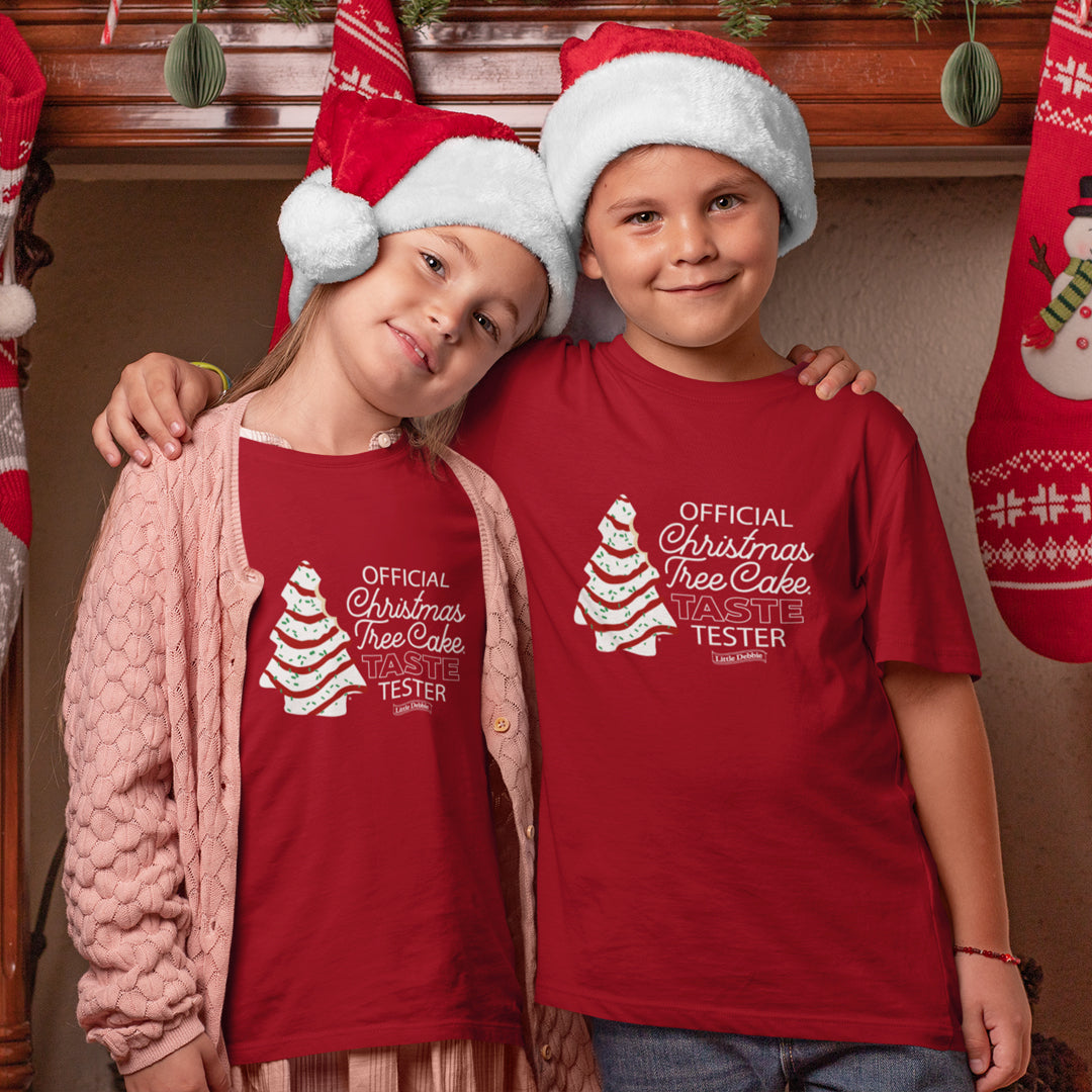 Two children wearing Santa hats are smiling and posing for the camera. The child on the right is wearing a red Little Debbie® Christmas Tree Cake Taste Tester Youth Shirt with the words 'OFFICIAL Christmas Tree Cake TASTE TESTER' printed in white alongside a graphic of a Christmas tree cake, with the Little Debbie® logo beneath. The child on the left is leaning on the right child's shoulder, wearing a similar red Little Debbie® Christmas Tree Cake Taste Tester Youth Shirt under a pink cardigan. 