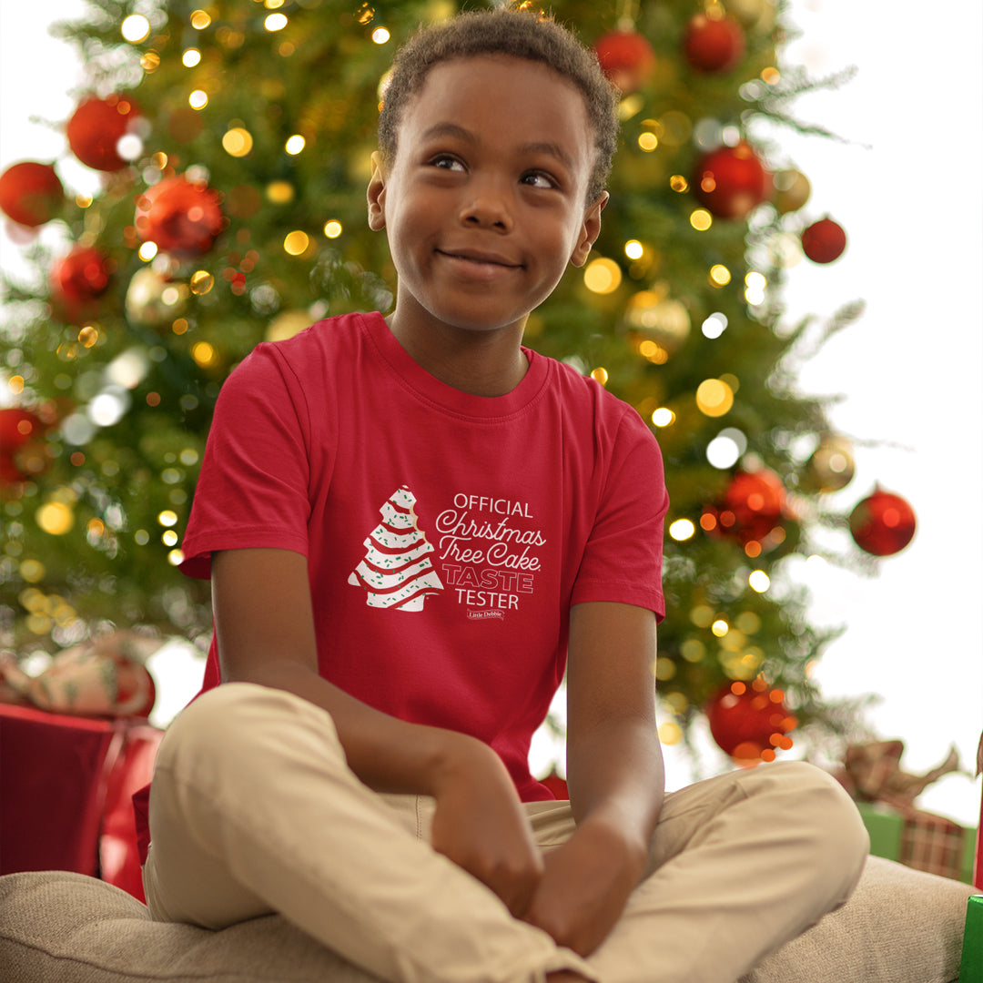 A young child with a joyful expression sits cross-legged in front of a decorated Christmas tree, wearing a red Little Debbie® Christmas Tree Cake Taste Tester Youth Shirt that reads 'OFFICIAL Christmas Tree Cake TASTE TESTER' with a graphic of a white Christmas Tree Cake and the Little Debbie® logo. The warm glow of the tree lights highlights the festive atmosphere.