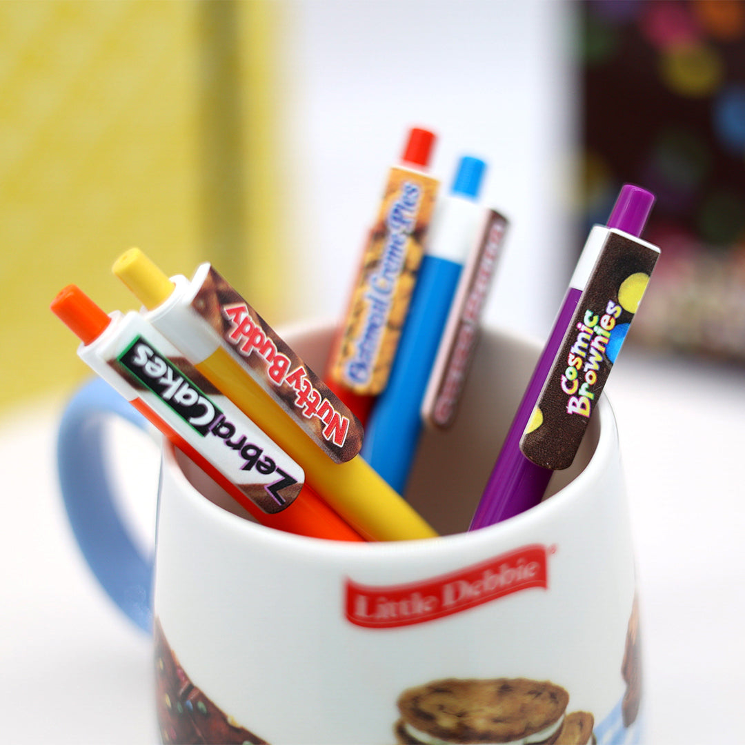 Five pens from the Little Debbie Scribbles collection, each representing a different Little Debbie treat, arranged in a Little Debbie mug featuring Cosmic Brownies, Oatmeal Creme Pie and Nutty Buddy..