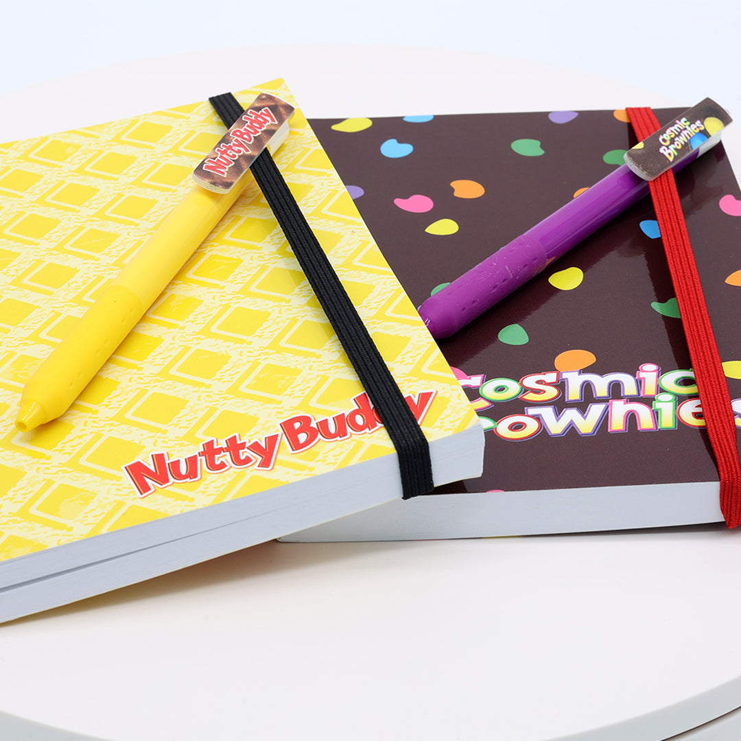 Nutty Buddy and Cosmic Brownie pens from the Little Debbie Scribbles set clipped into the strap of Nutty Buddy journal and a Csmic Brownie journal.