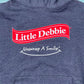 Close-up view of a blue Little Debbie Unwrap a Smile hoodie, featuring a large 'Little Debbie red ribbon logo in red with white lettering. Just below the logo, there's a white slogan that reads 'Unwrap A Smile' on the same blue background.