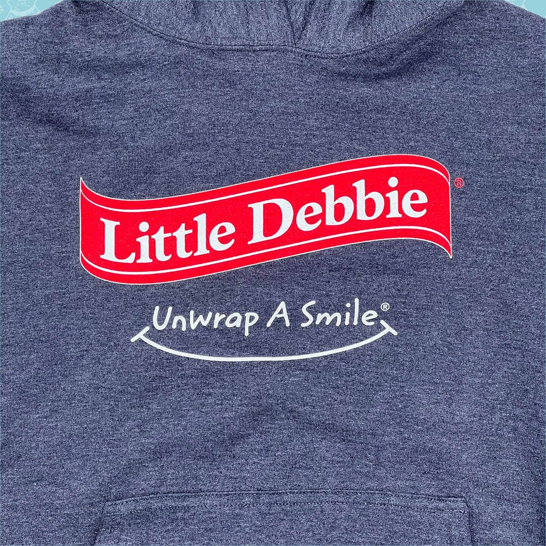 Close-up view of a blue Little Debbie Unwrap a Smile hoodie, featuring a large 'Little Debbie red ribbon logo in red with white lettering. Just below the logo, there's a white slogan that reads 'Unwrap A Smile' on the same blue background.