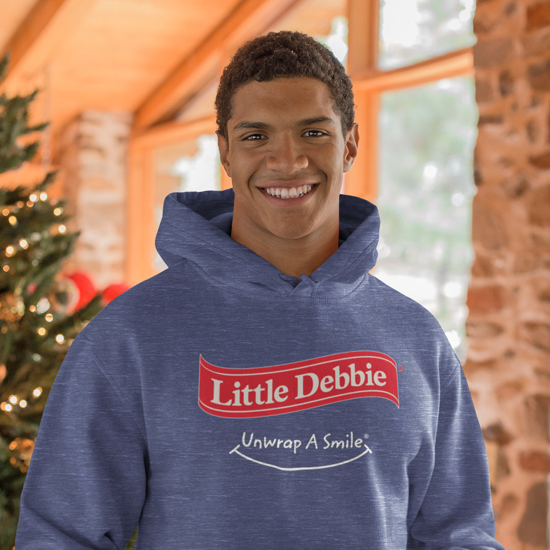 Man wearing a blue Little Debbie Unwrap a Smile hoodie with a Little Debbie red ribbon logo in red and white lettering on the front. The hoodie also displays the slogan 'Unwrap A Smile' below the logo. The man is smiling, standing indoors next to a decorated Christmas tree with a wooden and stone architectural background.