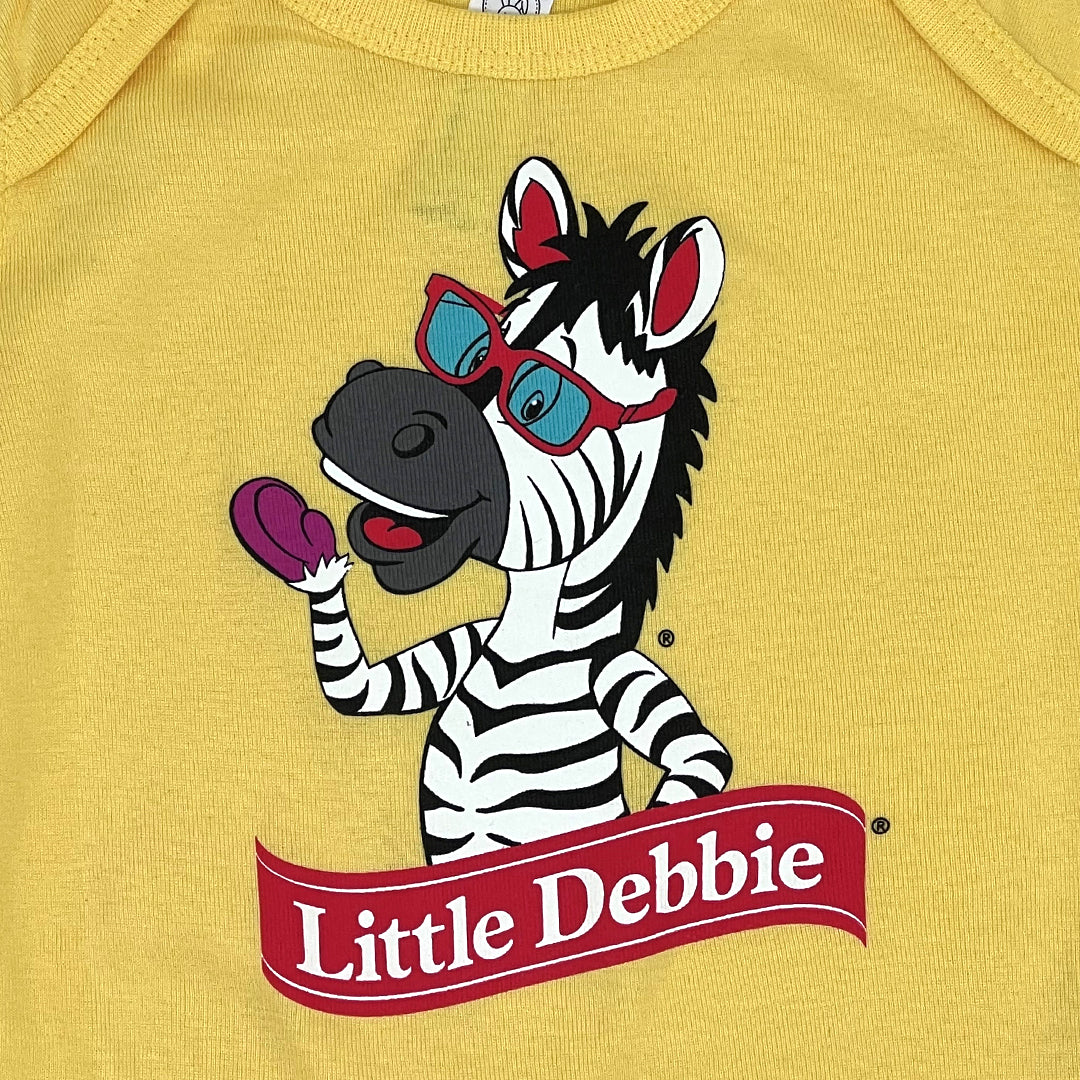 Detailed view of a yellow Little Debbie onesie, featuring a design of Zain the Zebra™ in a half-body image, waving. Zain appears to be positioned behind the Little Debbie Ribbon Logo.