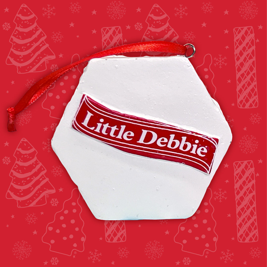 The back of the Little Debbie Zebra Cake ornament, displaying a white icing surface with the 'Little Debbie' logo across a red banner. The ornament is suspended by a shiny red ribbon and set against a red background decorated with white snowflakes and Christmas Tree Cake outlines.