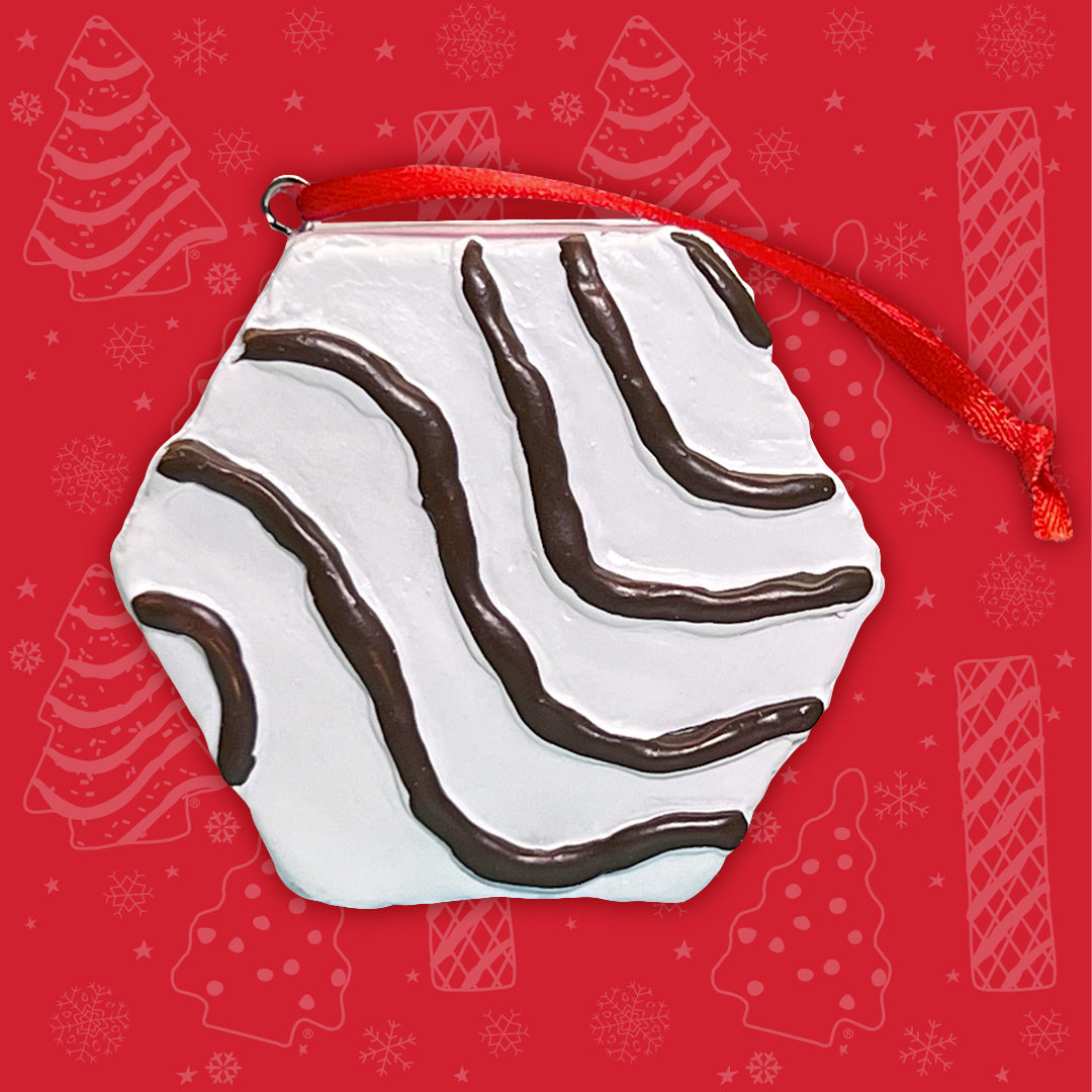 A ornament designed to resemble a Little Debbie Zebra Cake, with white icing and characteristic stripes, hangs by a red ribbon. It's presented against a festive red background with decorative snowflakes and Christmas Tree Cake silhouettes.