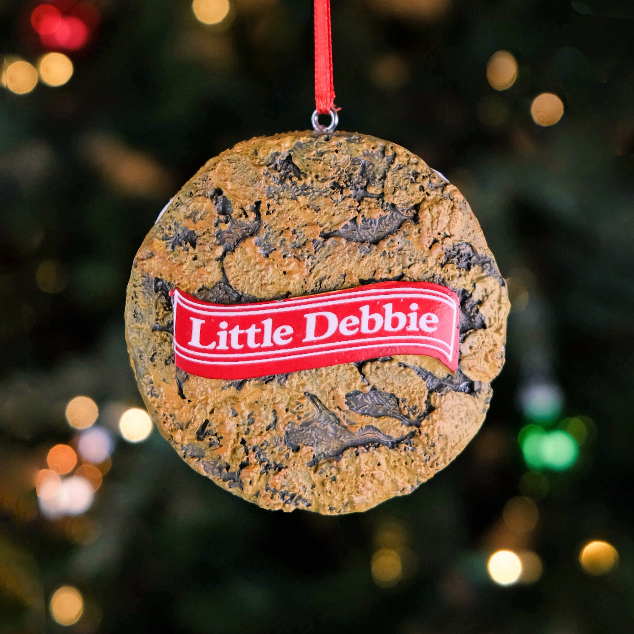 Back of the Little Debbie Oatmeal Creme Pie Ornament showing the textured cookie design with the red Little Debbie ribbon logo, hanging by a red ribbon in front of a softly lit Christmas tree.