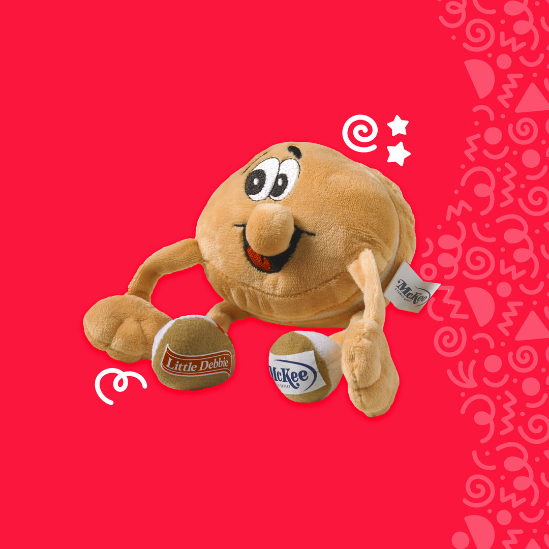 The image displays an Oatie plush toy against a vibrant red background with decorative white doodles. The plush is designed to resemble an Oatmeal Creme Pie with a smiling face, complete with eyes, a nose, and a cheerful mouth. It's depicted with arms and legs, giving it a lively and anthropomorphic charm. In one hand, it holds a miniature mug with the Little Debbie logo, while the other hand is raised as if waving. 