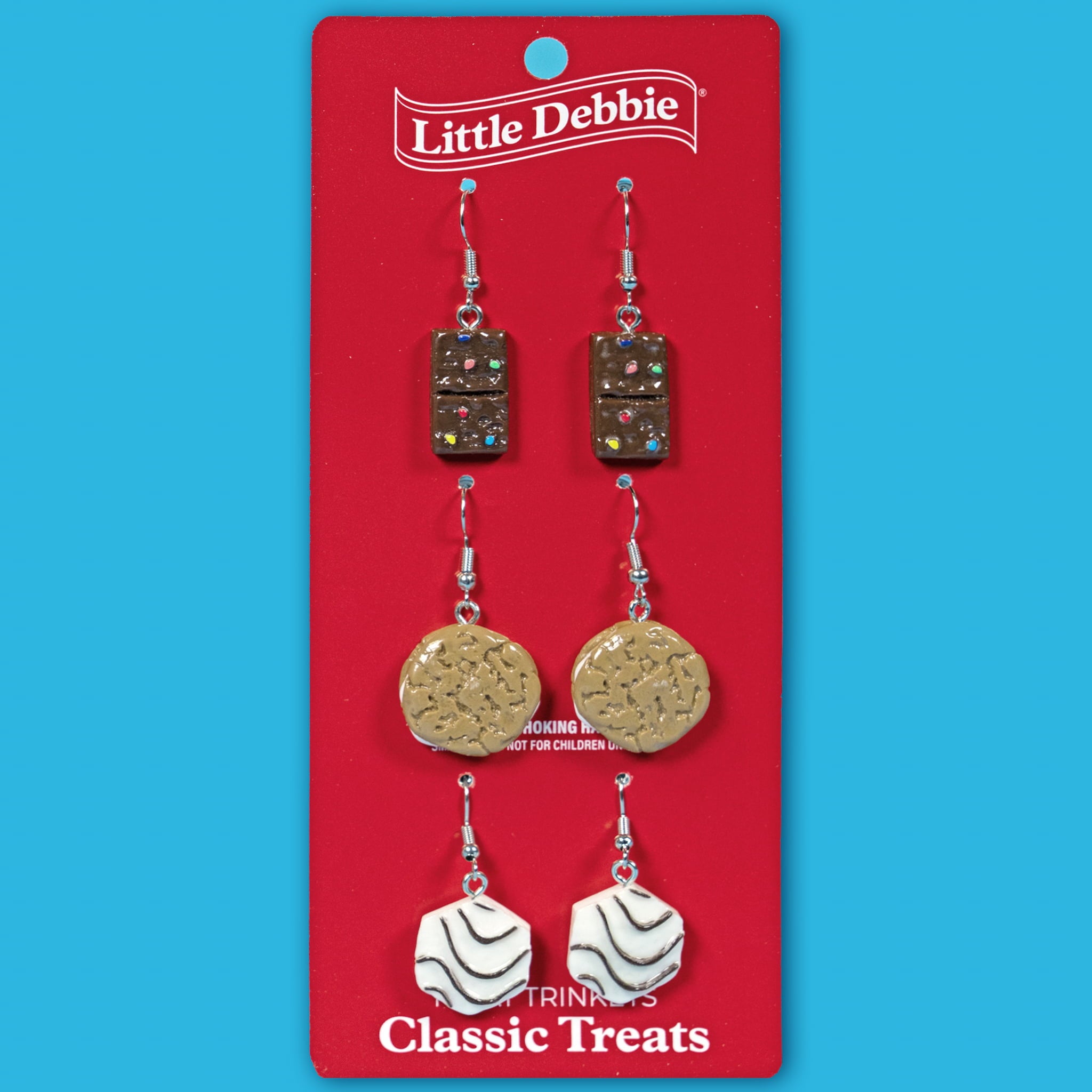 A red earring card with the Little Debbie logo displays the Classic Treats Earring Set, featuring three pairs of dangling earrings inspired by iconic snacks — Cosmic Brownies on top, Oatmeal Creme Pies in the middle, and Zebra Cakes on the bottom — against a bright blue background.